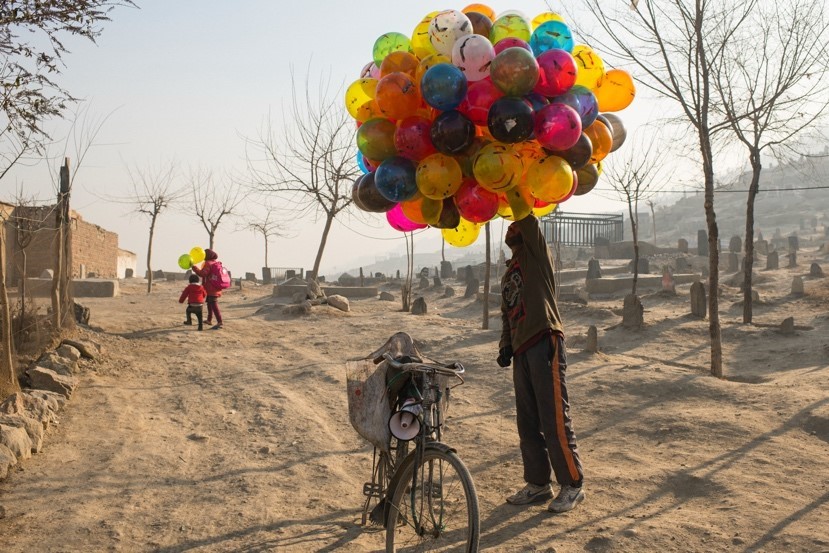A man standing on a sand covered street holding a large bundle of balloons standing next to a bicycle.