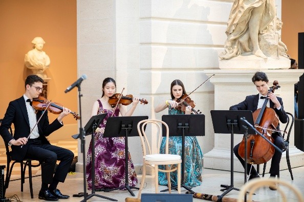 A string quartet playing their instruments with a sculpture of a woman in the background. 