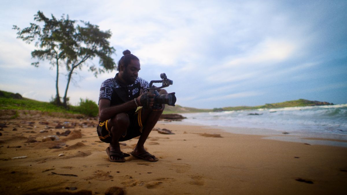 A man crouches on a beach with a film camera.