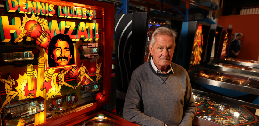 A man standing next to a pinball machine titled Dennis Lillee’s Howzat!.