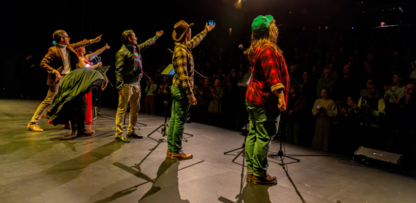 Six actors stand on a lit stage facing a seated crowd