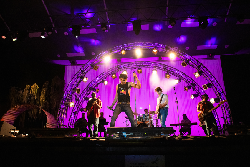 A group of people performing on a stage, holding instruments. A man holding a microphone is front and centre.