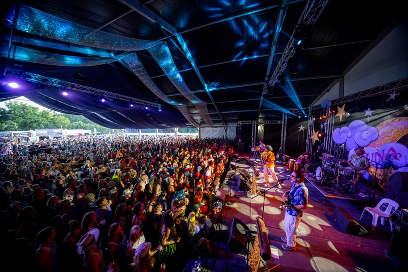A band plays on stage in front of a packed audience illuminated by colourful lights. 