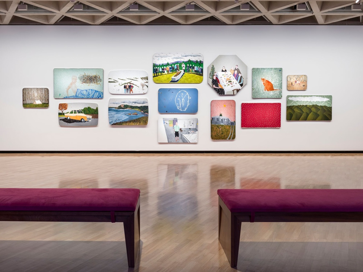 Installation view of a group of 14 paintings in varied shapes and sizes hanging in a salon-style arrangement on a white wall. The paintings feature landscapes, figures and abstract compositions. Two purple benches in the foreground are facing the installation.