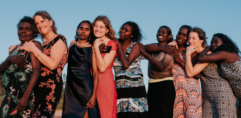 Nine women stand in a line against a blue sky backdrop, embracing each other.