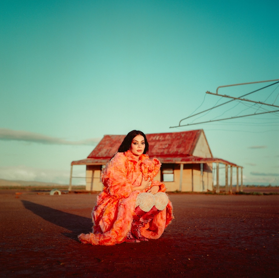 Australian singer Thelma Plum dressed in a vibrant orange dress and red boots crouched in front of a small homestead building next to a Hills Hoist clothesline surrounded by red dirt. 