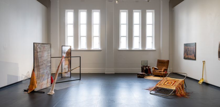 A gallery exhibition space with six narrow windows at one end and various artworks on display on the floor and walls.