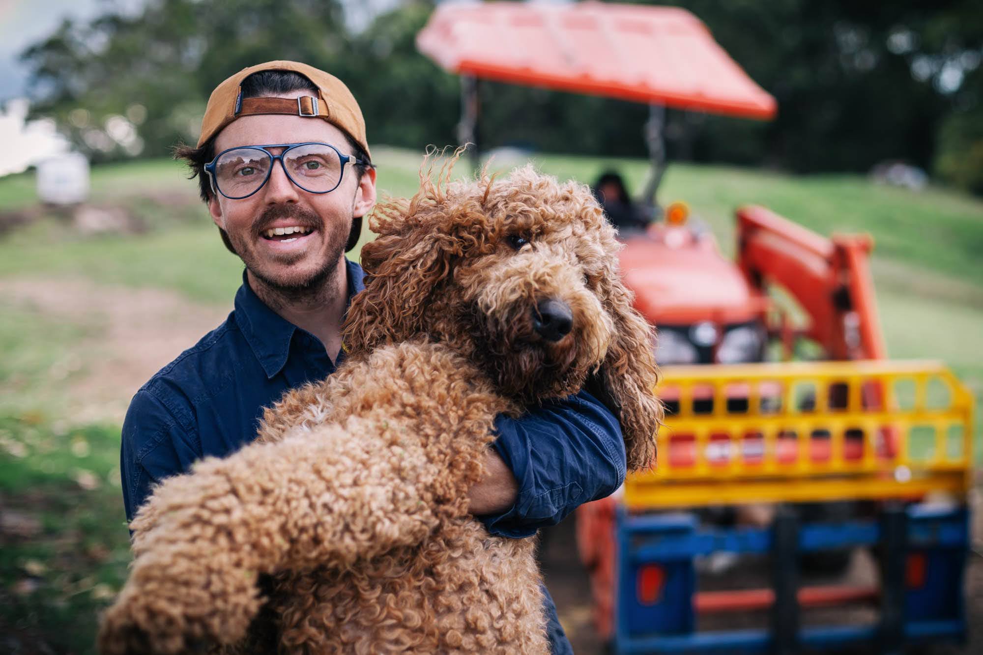 A Curious Tractor Co-founder Nicholas Marchesi. holding a large dog. A tractor and field is visible in the background.