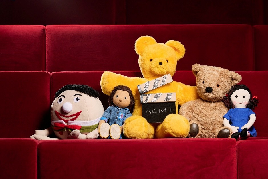 The toys from Play School sitting together in plush red cinema seats with a movie slate that reads ‘ACMI’. 