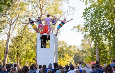 Circus performers holding hands while standing on top of a structure outside with trees in the background.
