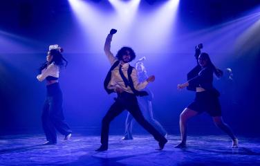 Four dancers performing on stage with blue lighting. One dancer is in the foreground and 3 in the background.