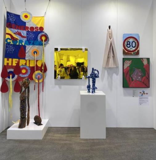 5 contemporary art pieces hang on a wall of a temporary gallery space. Two pieces are on plinths. On the wall there is hanging textile in primary colours, a square yellow painting showing people in a crowded room, a small  beige fabric piece, a square pai