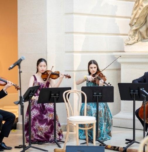 A string quartet playing their instruments with a sculpture of a woman in the background. 