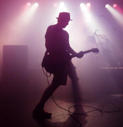 Musician stands on stage holding a guitar. Another guitarist is in the background. The stage is flooded with redish/pinkish light.