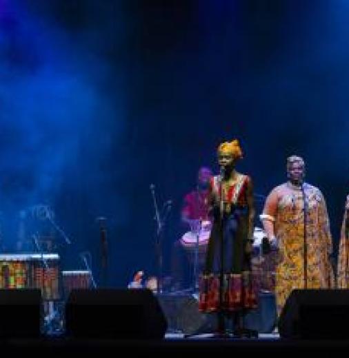 This image shows three singers in colourful, patterned clothing, with a guitarist and drummer, on a lit performance stage as part of the opening night of the 2021 Castlemaine State Festival. Ajak Kwai and Riel Dë Nyiër: World premiere at the Castlemaine 