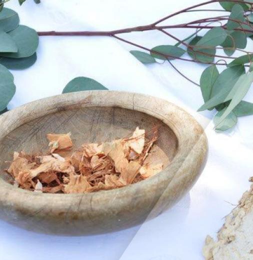 A wooden ceremonial bowl rests on a table with a white cloth. Gum tree leaves surround the bowl and a piece of bark rests to the right. The bowl contains bark fragments. 
