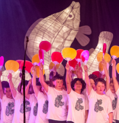 Primary school aged boys sing in a choir. They wear white t-shirts with black patterns and hold up paddles. A white fish sculpture is lit-up behind them. 
