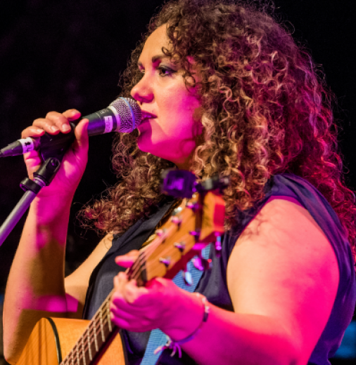 First Nations woman Deline Briscoe sings into microphone while performing on stage. She holds a guitar. 