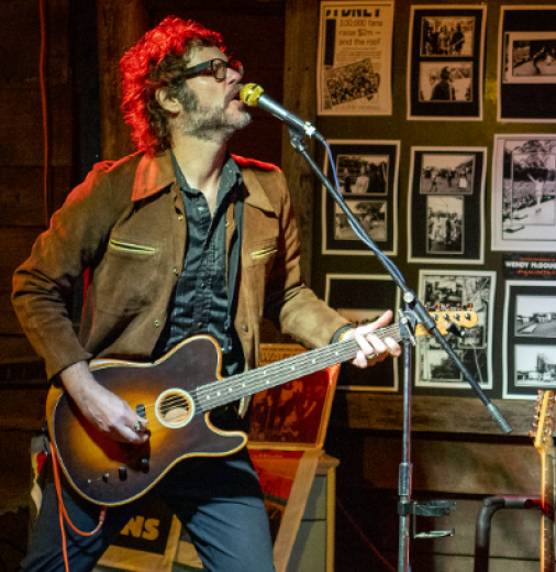 Man plays guitar in a pub venue singing into a microphone. A man and woman watch on.