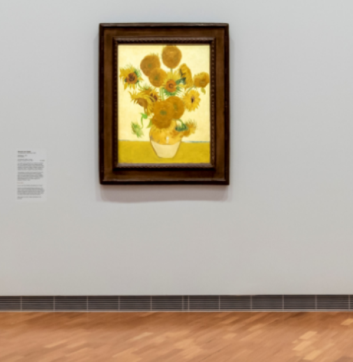 Painting of sunflowers in a vase. The painting is in a wooden frame, and is hanging on the grey gallery wall.