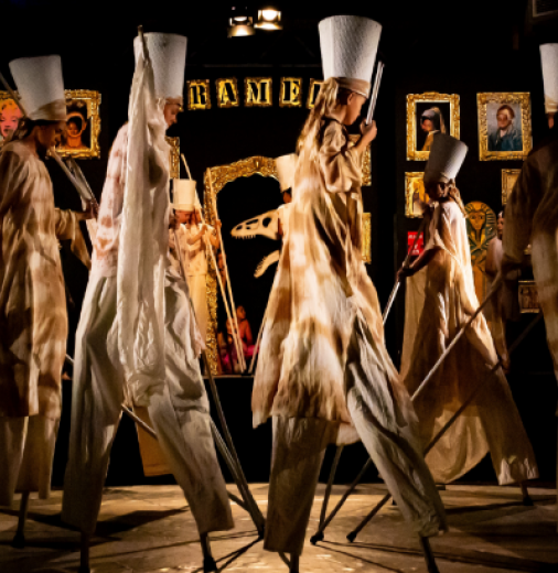 Circus performers walk on stilts on stage wearing long flowing white clothing and tall white hats