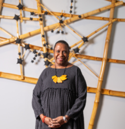 Artist, Gail Mabo, stands in front of artwork in a gallery. Artwork is made from timber and is adorned with black stars.