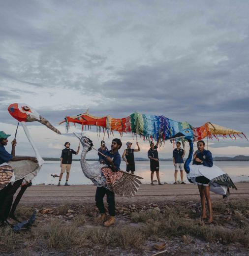 A group of people on a beach showcasing vibrant puppetry that features First Nations designs. 