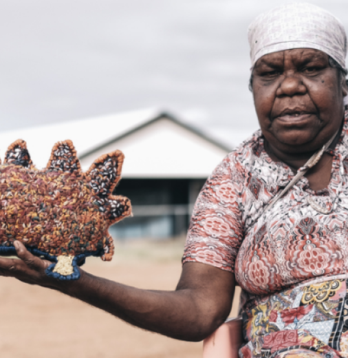 First Nations artist Marlene Rubuntja wears a dress with red, white, balck and yellow designs. She sits in a rural setting with a shed in the background, she is looking at the camera with one arm outstretched hold an echidna she stitched.