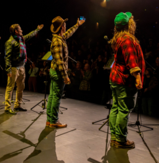 Six actors stand on a lit stage facing a seated crowd