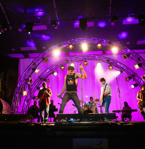 A group of people performing on a stage, holding instruments. A man holding a microphone is front and centre.