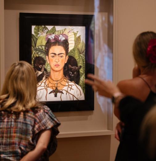 People in an exhibition looking at a painting of artist Frida Kahlo. 