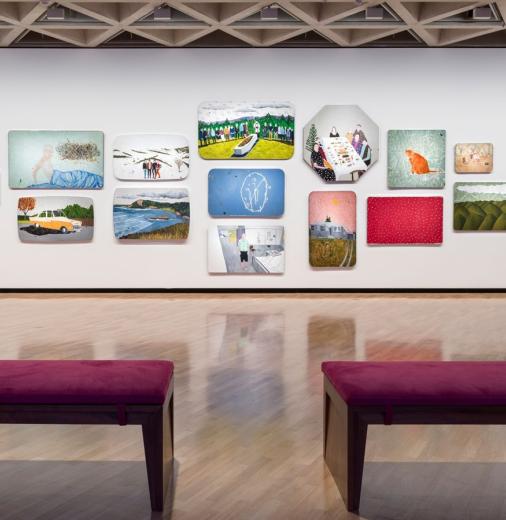 Installation view of a group of 14 paintings in varied shapes and sizes hanging in a salon-style arrangement on a white wall. The paintings feature landscapes, figures and abstract compositions. Two purple benches in the foreground are facing the installation.