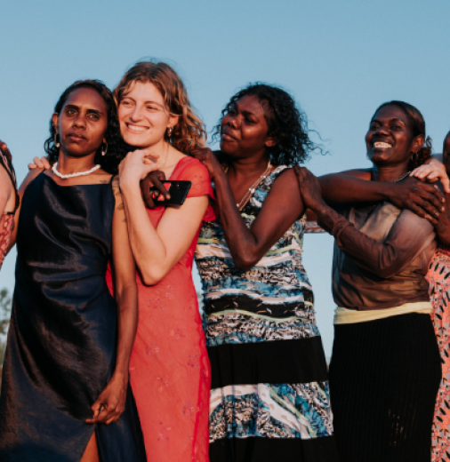 Nine women stand in a line against a blue sky backdrop, embracing each other.