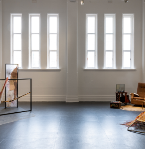A gallery exhibition space with six narrow windows at one end and various artworks on display on the floor and walls.