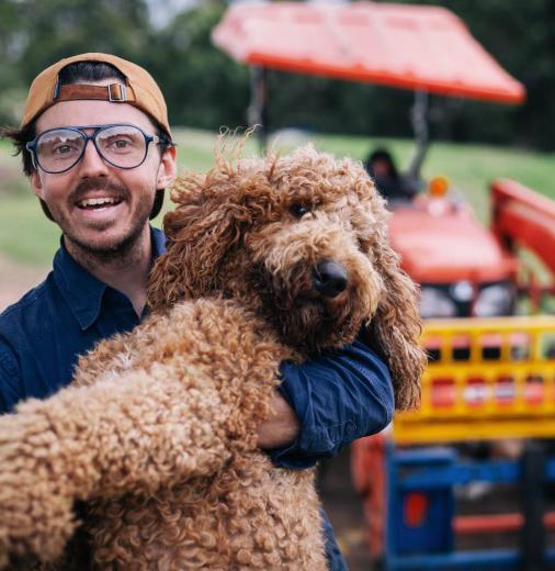 A Curious Tractor Co-founder Nicholas Marchesi. holding a large dog. A tractor and field is visible in the background.