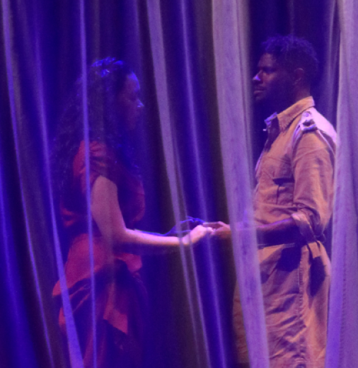 First Nations male actor holds hands with a female actor on stage during a performance. Sheer curtains surround them and the stage is lit in purple lights.