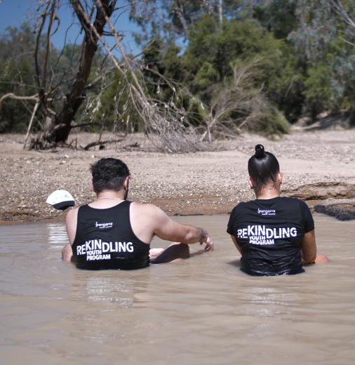 Male and two female youth sitting in a river, wearing t-shirts reading ‘Rekindling Youth Program’