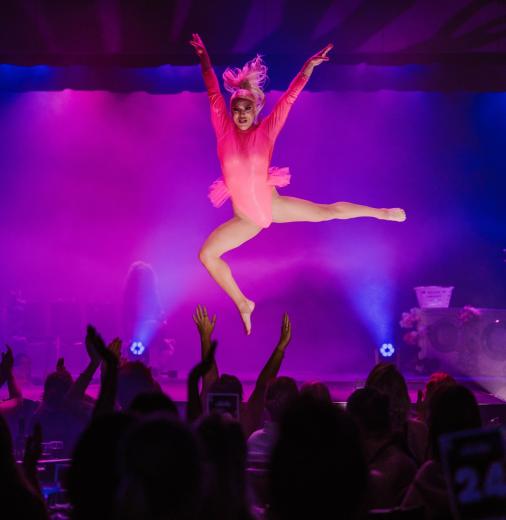 Performer in pink leotard is suspended mid-air with arms and legs outstretched. Crowd below applauds.