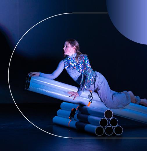 A performing artist dressed in purple clothing rests on a prop made of piping, and is illuminated by a spotlight.