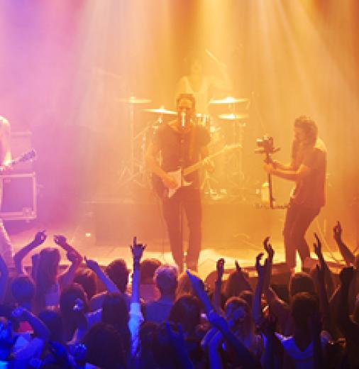  A rock band performs live on stage, illuminated by vibrant, colourful stage lights. A cheering crowd, with hands raised, enjoys the energetic performance.