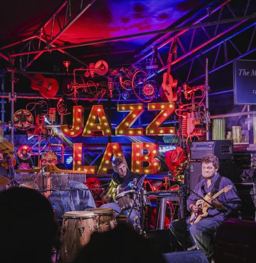 A band plays their instruments in front of a lit-up sign that reads ‘Jazz Lab’. 
