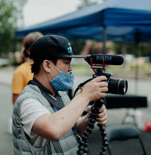 A screen worker on set holding a video camera with microphone and tripod. They are wearing a baseball cap and facemask.