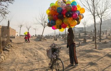 A man standing on a sand covered street holding a large bundle of balloons standing next to a bicycle.