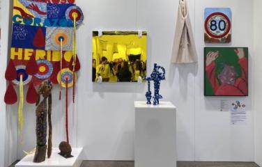 5 contemporary art pieces hang on a wall of a temporary gallery space. Two pieces are on plinths. On the wall there is hanging textile in primary colours, a square yellow painting showing people in a crowded room, a small  beige fabric piece, a square pai