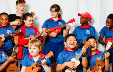 A group of primary school children play instruments in their school classroom. They are smiling and laughing.