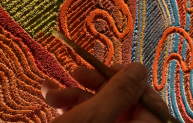Close up of a hand holding a paintbrush against a brightly-coloured artwork