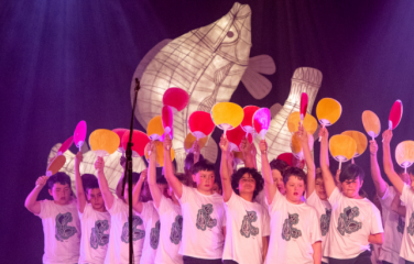 Primary school aged boys sing in a choir. They wear white t-shirts with black patterns and hold up paddles. A white fish sculpture is lit-up behind them. 