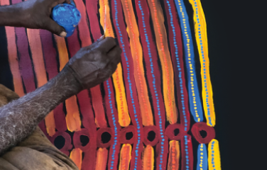 An artist's hands, holding a paint jar and painting tool, working on a colourful Indigenous artwork, which features yellow, blue, orange and red designs. 