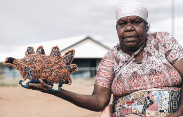 First Nations artist Marlene Rubuntja wears a dress with red, white, balck and yellow designs. She sits in a rural setting with a shed in the background, she is looking at the camera with one arm outstretched hold an echidna she stitched.