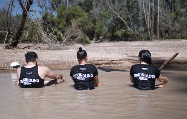 Male and two female youth sitting in a river, wearing t-shirts reading ‘Rekindling Youth Program’